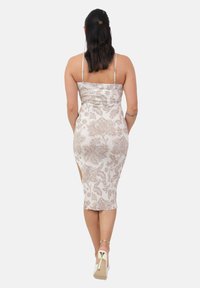 Donna con capelli scuri in un vestito a fantasia floreale beige e tacchi alti bianchi, che si allontana su uno sfondo chiaro e semplice.