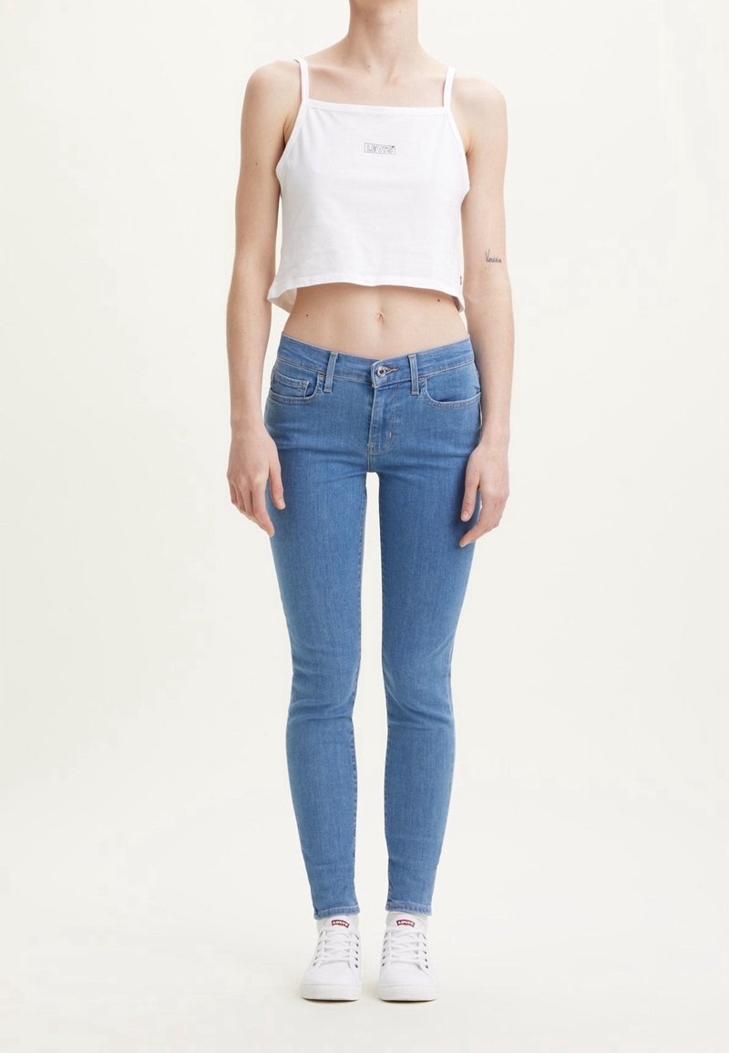 Bílý zkrácený top bez rukávů s tenkými ramínky, modré skinny džíny a bílé tenisky. Outfit má jednoduchý design a minimalistické detaily.
