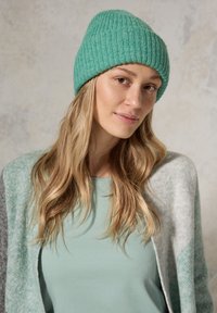 Ribbede grønne strikhætte, blød struktur. Model iført en mintgrøn top, med en lys grå og teal cardigan med geometrisk mønster.