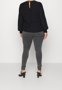 Blouse noire à manches longues transparentes, détail dos avec ouverture en forme de goutte, portée avec un jean skinny gris et des sandales à talons blocs noires.