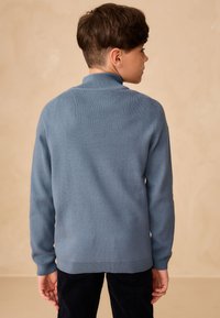 Maglione blu a maglia con colletto alto, caratterizzato da polsini e orlo a coste, indossato con pantaloni scuri. La texture appare liscia e aderente.