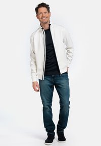 Witte bomberjack met hoge kraag, zwarte rits en zijstrip, gedragen over een zwart shirt en blauwe jeans, gecombineerd met zwarte sneakers.