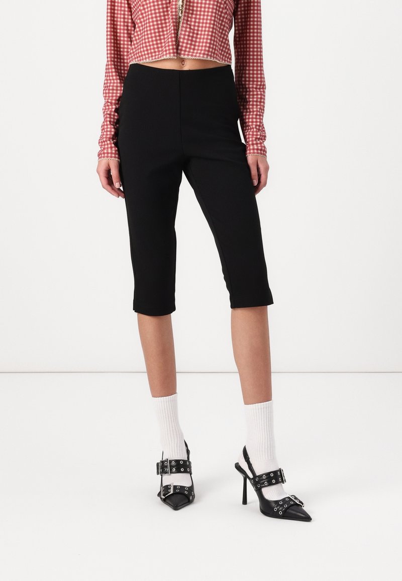Femme portant un pantalon noir ajusté longueur genou, des chaussettes blanches, des chaussures noires à talons hauts avec boucles, et un top court à carreaux rouges.