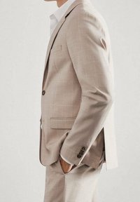 Lichtbeige geruit pak met een getailleerd ontwerp, een enkelsluitend colbert, zijzakken, een notched revers en bruine knopen. Witte blouse eronder.