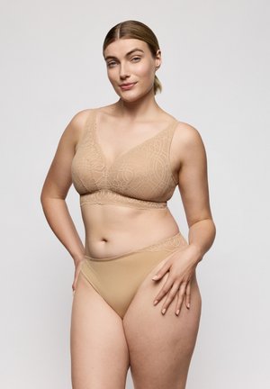 Vrouw met blond haar, gekleed in een nude kanten bralette en bijpassend hoog gesneden ondergoed, staat tegen een eenvoudige lichte achtergrond en glimlacht zelfverzekerd.