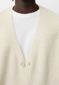 Beige ullkofta med djup V-ringning, med en åldersknappstängning och ribbad textur. En vit T-shirt syns under.