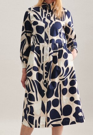 Beżowa sukienka z długim rękawem i zapięciem na guziki, z dużymi granatowymi abstrakcyjnymi wzorami liści i kropek, kołnierzykiem i bocznymi kieszeniami, noszona przez kobietę.