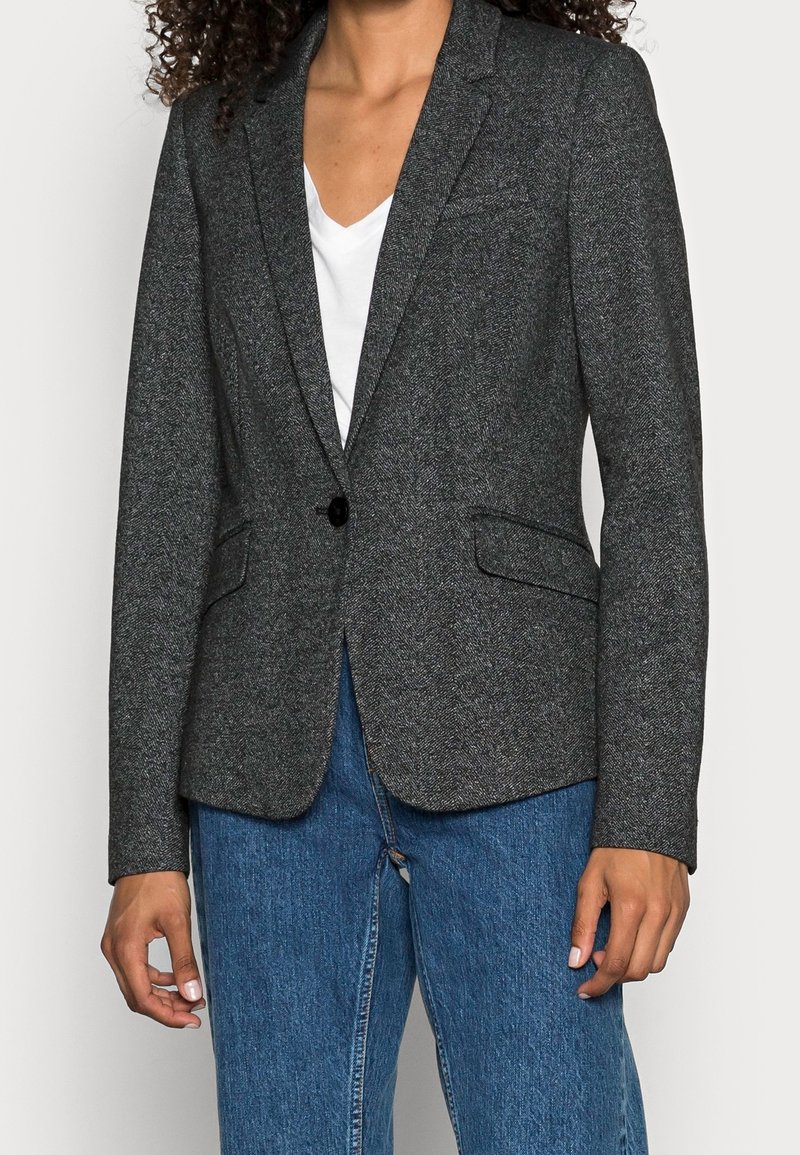 Personne portant un blazer gris foncé à un bouton sur un t-shirt blanc col en V et un jean bleu, les mains détendues le long du corps.