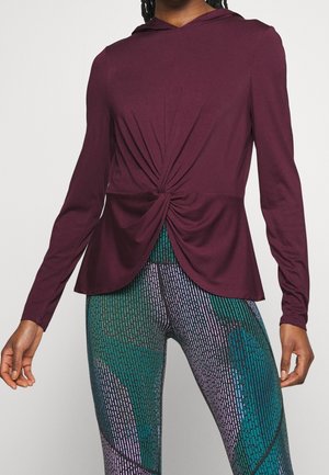Vrouw draagt een lange mouwen maroon top met gedraaide voorkant en hoog getailleerde legging met een abstract patroon in groen, paars en zwart.