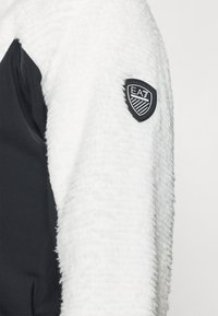 Valge fliisist jakk, mille ühel küljel on must sile kangas. Disainile lisab detaili must EA7 logo plaastriga varrukal.