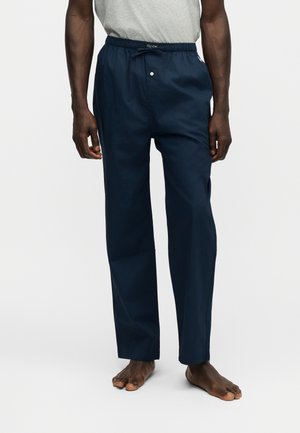 Pantalon bleu foncé à cordon porté par une personne pieds nus se tenant devant un fond clair uni, associé à une chemise gris clair.