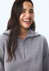 Grauer Zip-Up-Pullover mit einem gerippten Kragen und einem silbernen Reißverschluss. Weiches, strukturiertes Material, lockere Passform und minimale Nähte.