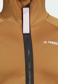 Veste Adidas Terrex marron avec fermeture éclair noire et languette blanche au centre devant.