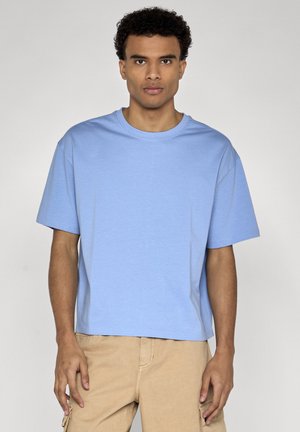 Ung mand med kort krøllet hår iført en lyseblå oversized T-shirt og beige cargobukser mod en ensfarvet baggrund.