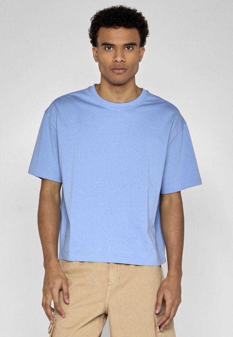 Jeune homme aux cheveux courts et bouclés portant un T-shirt oversize bleu clair et un pantalon cargo beige sur un fond uni.
