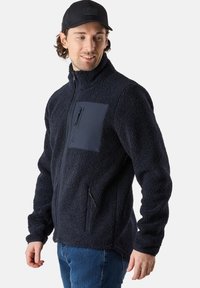 Giacca in pile di un blu scuro, con colletto alto, chiusura zip sul davanti e tasca sul petto. Texture morbida con dettagli in tessuto a contrasto.