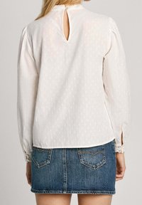 Eine Frau trägt eine weiße Bluse mit langen Ärmeln und Spitzenmanschetten sowie einem Schlüsselverschluss auf der Rückseite, kombiniert mit einem blauen Jeans-Minirock.