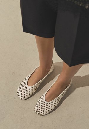Pieds portant des chaussures blanches à motifs en treillis, sans lacets, avec un pantalon noir raccourci, debout sur une surface beige clair.