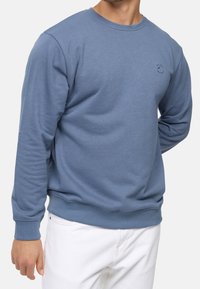 Homme portant un sweat-shirt bleu à manches longues avec des poignets côtelés et un pantalon blanc, la main relaxée à ses côtés, sur un fond uni.