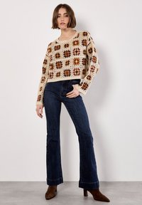 Crochet sweater with a beige background and colorful square patterns, paired with dark blue flared jeans and brown heeled boots.