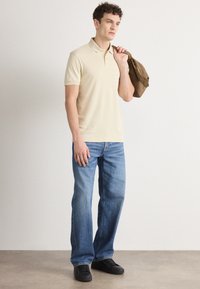 Beige pólóing klasszikus gallérral, világoskék egyenes szabású farmernadrággal és fekete sportcipőkkel, barna táskával a kezében.