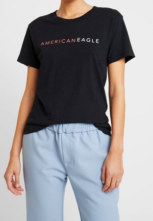 T-shirt en coton noir avec "AMERICAN EAGLE" imprimé en blanc et rouge. Design à col rond, manches courtes, associé à un pantalon bleu clair.