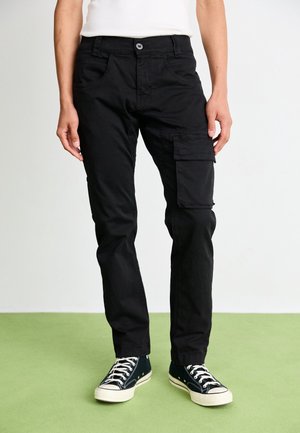 Person trägt schwarze Cargohose mit Klapptasche an einem Bein, weißes Hemd und schwarze Low-Top-Sneaker auf grünem Boden.