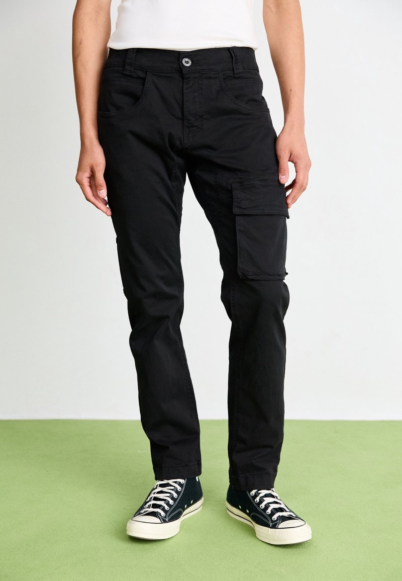 Personne portant un pantalon cargo noir avec une poche rabat sur une jambe, une chemise blanche et des baskets noires basses sur un sol vert.