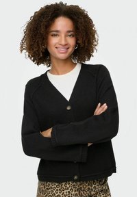 Cardigan nero con scollo a V, chiusura a un bottone e polsini a coste. Indossato sopra una maglietta bianca e abbinato a un pantalone con stampa leopardata.