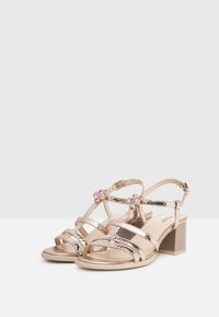 Sandali aperti con cinturini metallici dorati, tacco a blocco e fiori decorativi sulla parte anteriore. Texture liscia e design incrociato.