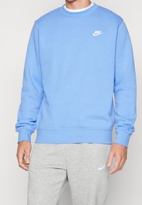 Sudadera azul claro con escote redondo, puños y dobladillo de canalé; con un logo blanco de Nike en el lado izquierdo del pecho. Combinada con pantalones de chándal grises.