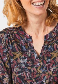 Blumenbluse mit dunklem Hintergrund und bunten Blattmustern in Rot-, Grün- und Goldtönen. V-Ausschnitt und geraffte Details.