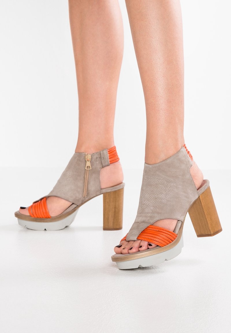 Jambes portant des sandales à talons ouverts, taupe et orange, avec des talons blocs en bois et des fermetures éclair latérales, debout sur un sol blanc.