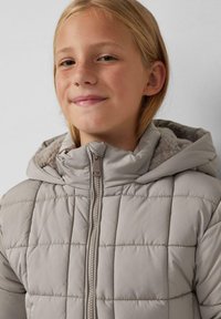 Nasmejana dijeca s laganom kosom nosi sivo izolirano jaknu s kapuljačom obloženom mekanim krznom, na jednoličnoj pozadini.