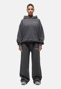 Sweat à capuche gris surdimensionné avec une poche avant et un logo blanc, associé à un pantalon de survêtement gris à jambes larges. Fabriqué en tissu doux et texturé.