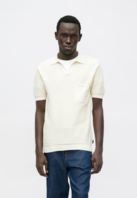 Κρεμ κοντομάνικο πόλο μπλουζάκι με υφασμένο μοτίβο, λαιμόκοψη σε σχήμα V και τσέπη στο στήθος, συνδυασμένο με σκούρα μπλε τζιν παντελόνια.