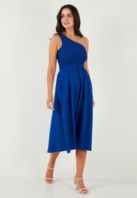 Blue one-shoulder dress with smocked bodice, flowing skirt, knee-length hem, and textured fabric. Worn with strappy heels.
