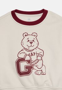 Creme sweatshirt med en maroon ribstrikket krave, der har en tegneserie-bjørn iført en sweater med "GAP," samt et stort maroon "G" grafik.