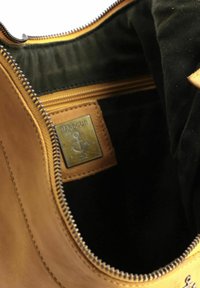 Borsa in pelle madreperlacea aperta che mostra un interno in tessuto nero con una tasca con zip e una placca metallica etichettata "Harbour" con un logo ad ancora.