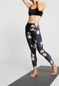 Zwarte bloemenleggings met roze en witte bloempatronen, hoge taille en een gladde, rekbare stof. Model staat blootsvoets op een mat.