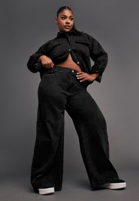 Mustapellinen farkkutakki, jossa on kaksi rintataskua, yhdistettynä korkeavyötäröisiin mustiin leveälahkeisiin farkkuihin. Sileä pinta, nappikiinnitys ja rento istuvuus.