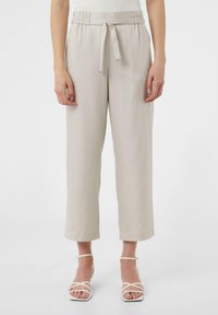 Jasno beżowe, luźne spodnie z pasem na sznurku, szerokimi nogawkami i gładką fakturą. Połączone z białymi sandałami na paski.