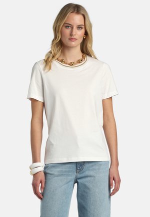 Femme blonde portant un t-shirt blanc uni à manches courtes, un jean bleu clair, un collier chaîne en or, des boucles d'oreilles en or et des bracelets blancs sur un fond blanc.