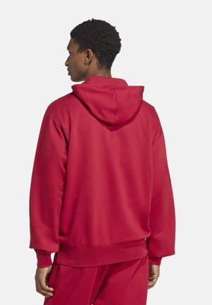 Homme de profil portant un sweat à capuche rouge uni et un pantalon rouge assorti sur un fond blanc.