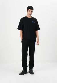 Camiseta negra de gran tamaño con texto azul, combinada con pantalones negros ajustados. La modelo lleva zapatos negros tipo slip-on. Diseño minimalista y colores monocromáticos.