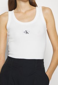 Vit ribbad linne med rund halsringning och breda axelband, med en Calvin Klein Jeans-logotyp i mitten. Kombineras med svarta byxor.