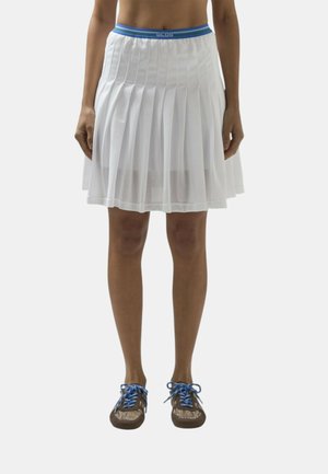 Donna con gonna bianca plissettata con fascia in vita blu e sneakers marroni con lacci blu, in piedi davanti a uno sfondo semplice.