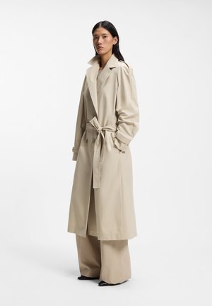 Mujer con un abrigo trench beige con cinturón, pantalones anchos beige y zapatos negros puntiagudos, de pie contra un fondo blanco.
