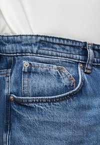 Tasca di jeans in denim blu con cuciture visibili, rivetti e passante per cintura su sfondo di tessuto bianco.