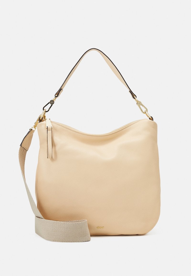 Sac à main en cuir beige avec bandoulière large en tissu détachable, petite poignée en cuir et ferrures dorées sur fond blanc.
